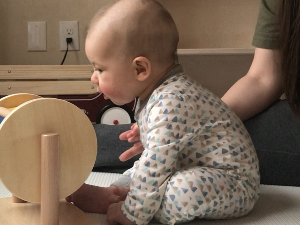 A baby sits with his hands on the ground watching a colorful spinning drum