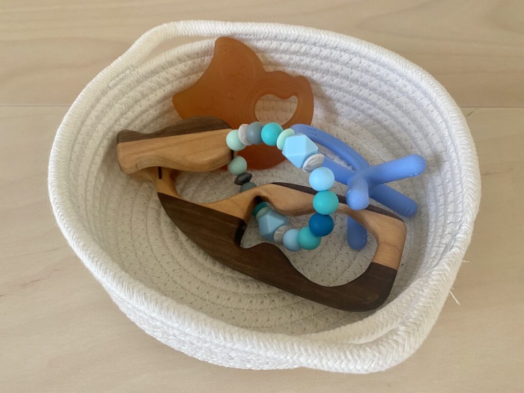 A cotton basket containing 3 natural teethers made of wood, rubber, and silicon.